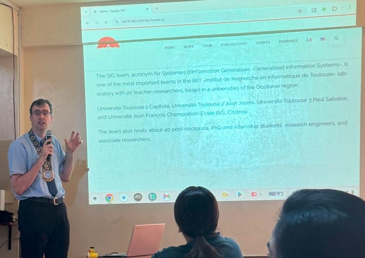 Philippe Roussille, standing, a microphone in one hand and the other raised, addresses the audience. Behind him, a projector displays the English presentation page of the SIG team at IRIT on the wall. In the foreground, two people seated with their backs to the camera follow the talk, and a yellow rubber duck sits on the desk beside a laptop.